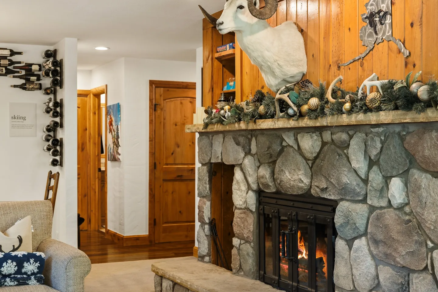Living room featuring a lit stone fireplace with wood paneled wall, ram taxidermy mount, and a hallway with wine racks.