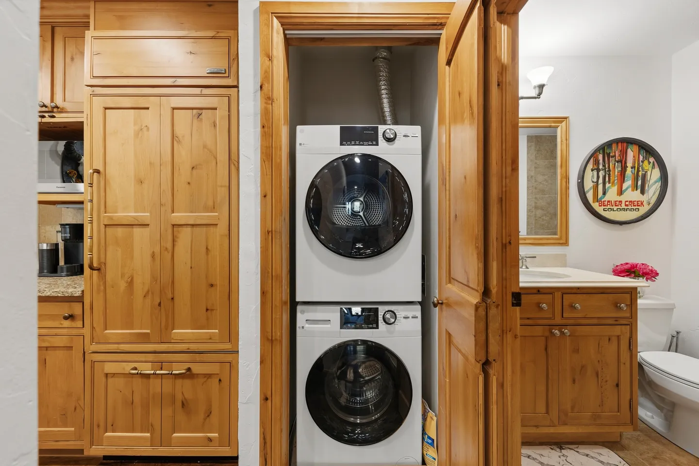A stacked white front-loading washer and dryer inside a wooden door closet adjacent to a bathroom and kitchen cabinetry.