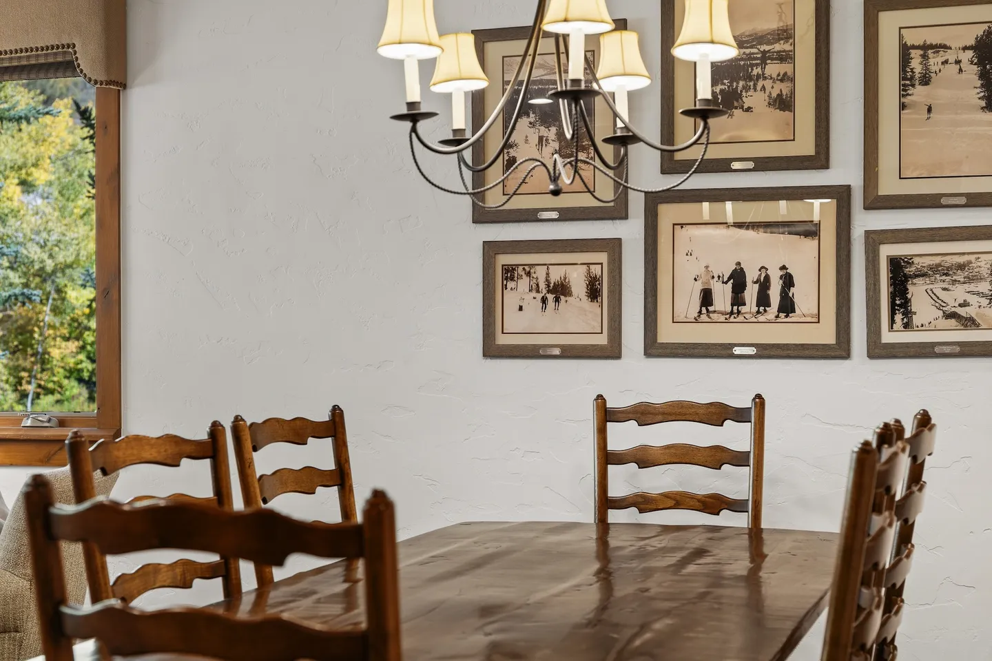 Dining area featuring a wooden table with matching ladderback chairs beneath a chandelier, with framed vintage winter photos on the wall.