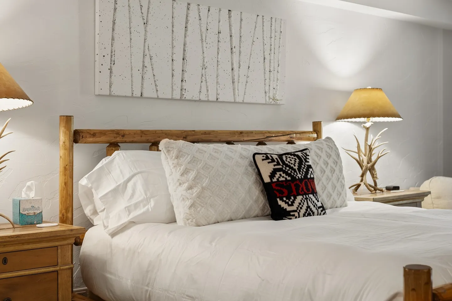 Close-up of a bed with a log headboard, white linens, an accent pillow, and nightstands with antler lamps.