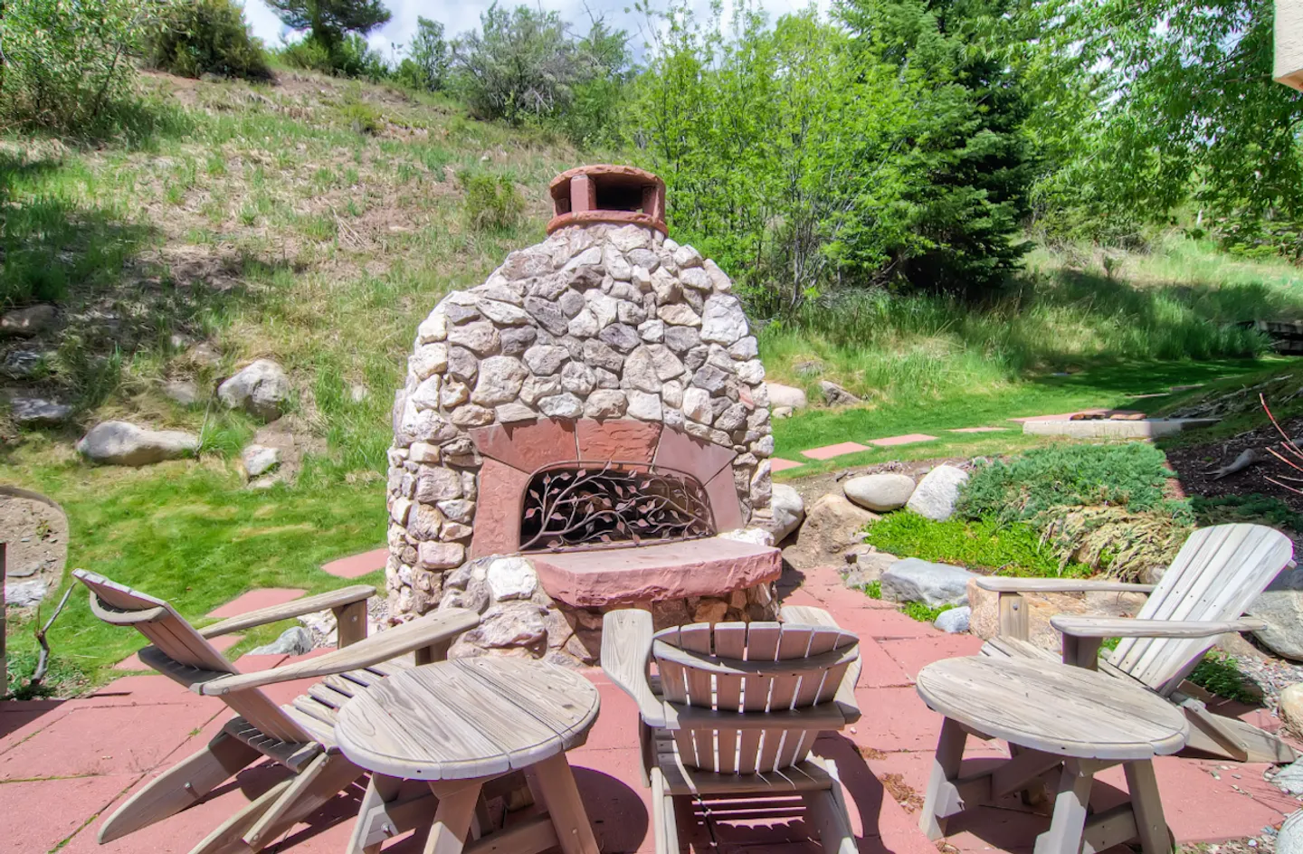 Outdoor stone fireplace with Adirondack chairs on a red paved patio surrounded by a grassy hillside.