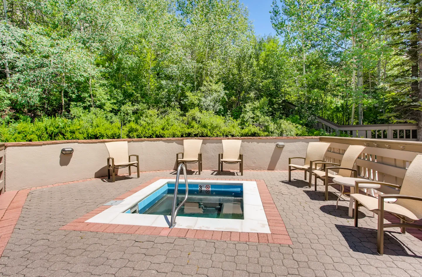 Outdoor hot tub embedded in a paved patio surrounded by seating and lush green trees.