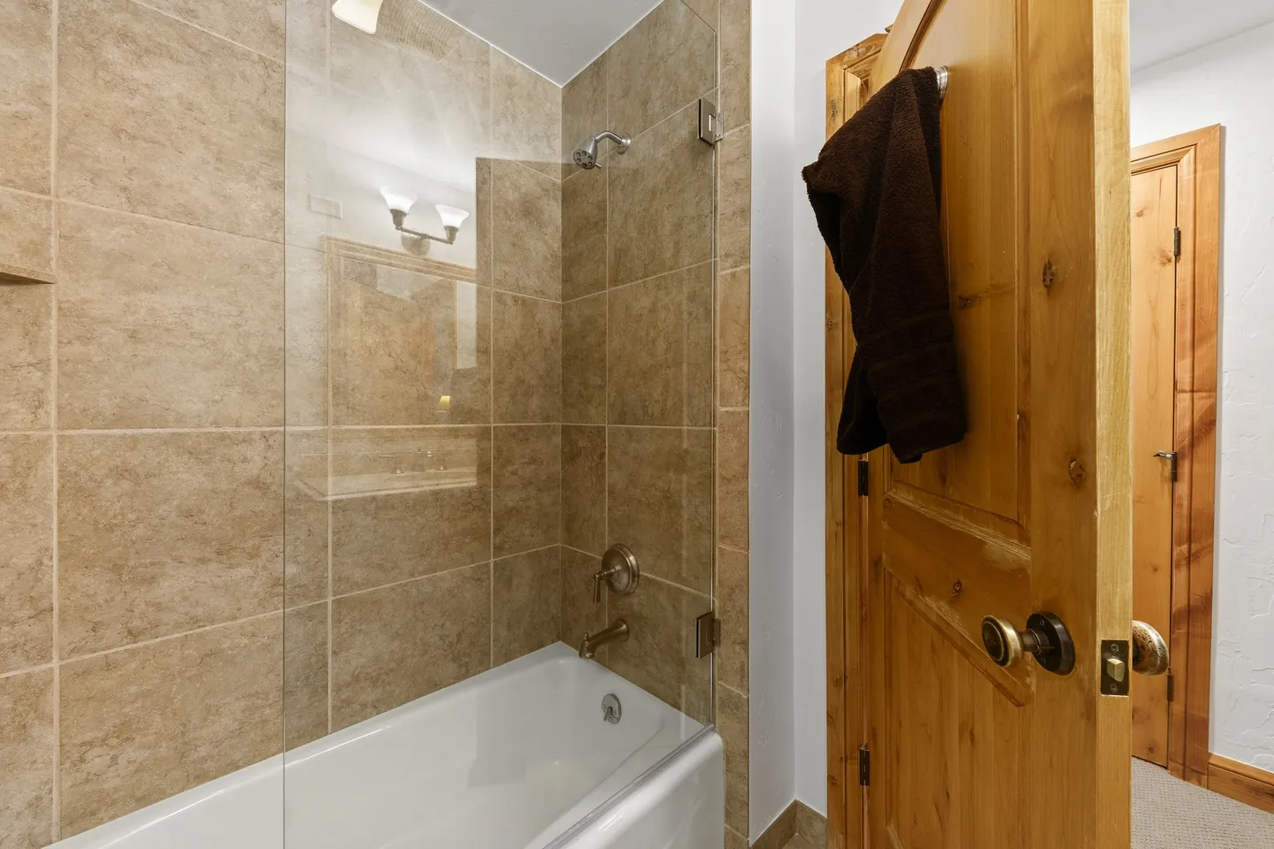 Bathroom showing a bathtub and shower combination with tan tile surround, a clear glass partition, and a wooden door.