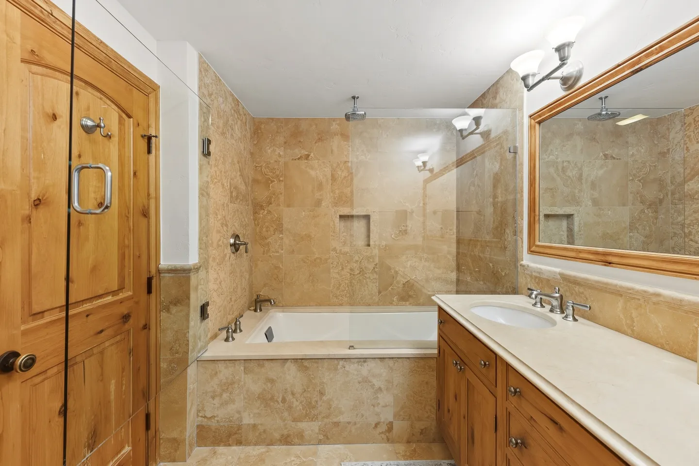 Bathroom with a drop-in bathtub surrounded by tan tile, a glass shower partition, and a matching wooden vanity and door.