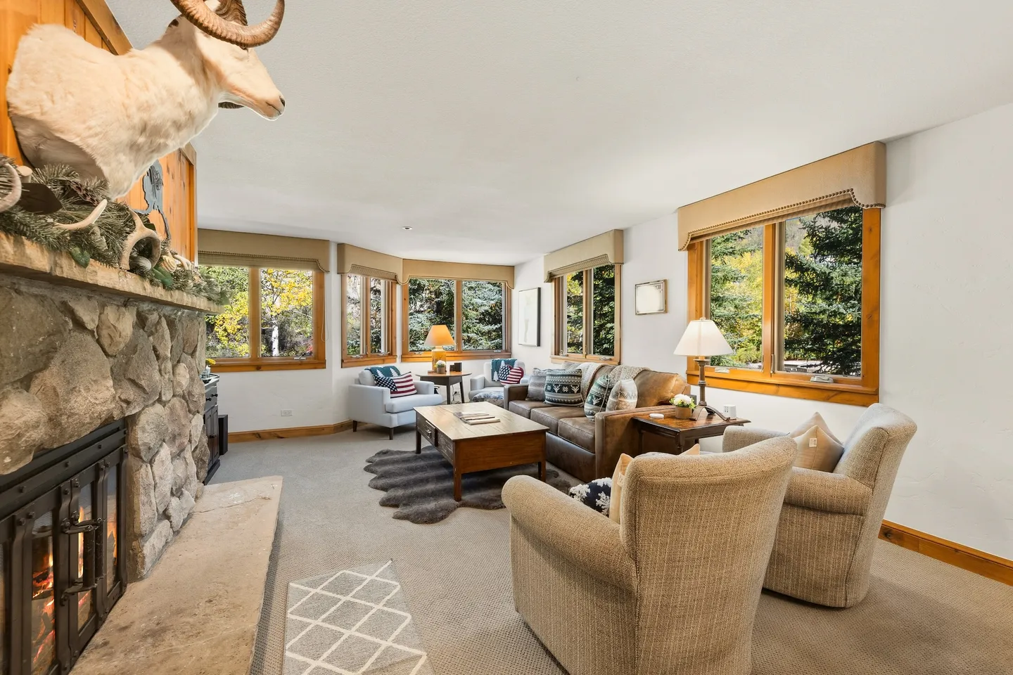 Living room with carpeted floor, stone fireplace, leather sofa, armchairs, and windows showing trees outside.