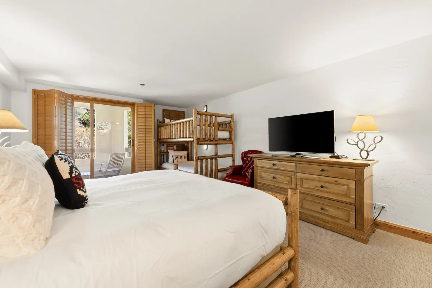 Bedroom featuring a wooden bed in the foreground, wooden bunk beds, a dresser with a television, and sliding glass doors with wooden shutters leading outside.
