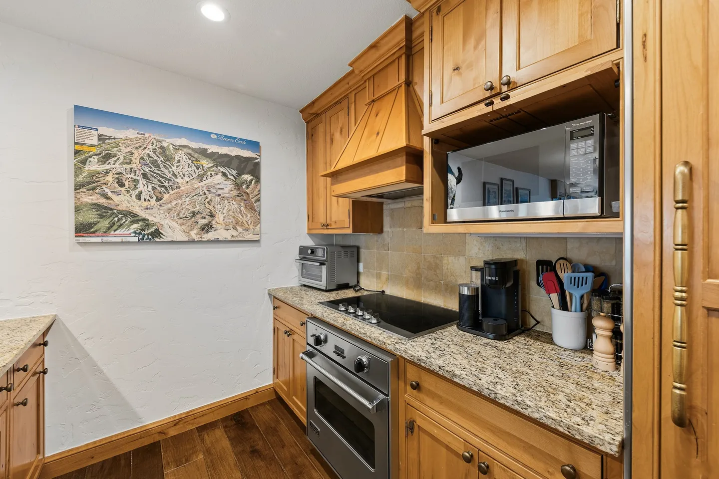 Kitchen with natural wood cabinets, granite countertops, an oven, smooth cooktop, and a ski trail map on the wall.