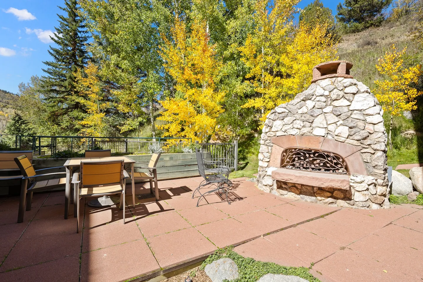 Outdoor patio with a stone fireplace, dining table, chairs, and vibrant yellow autumn trees in the background.