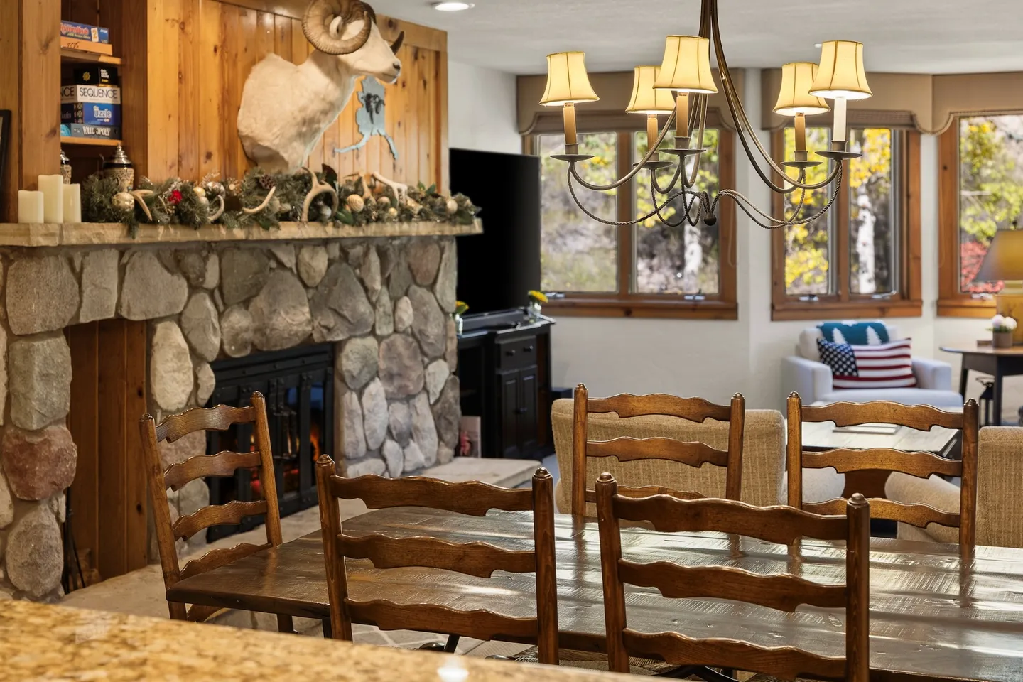 Dining area with a rustic wooden table and chairs in front of a stone fireplace with a mounted bighorn sheep head.