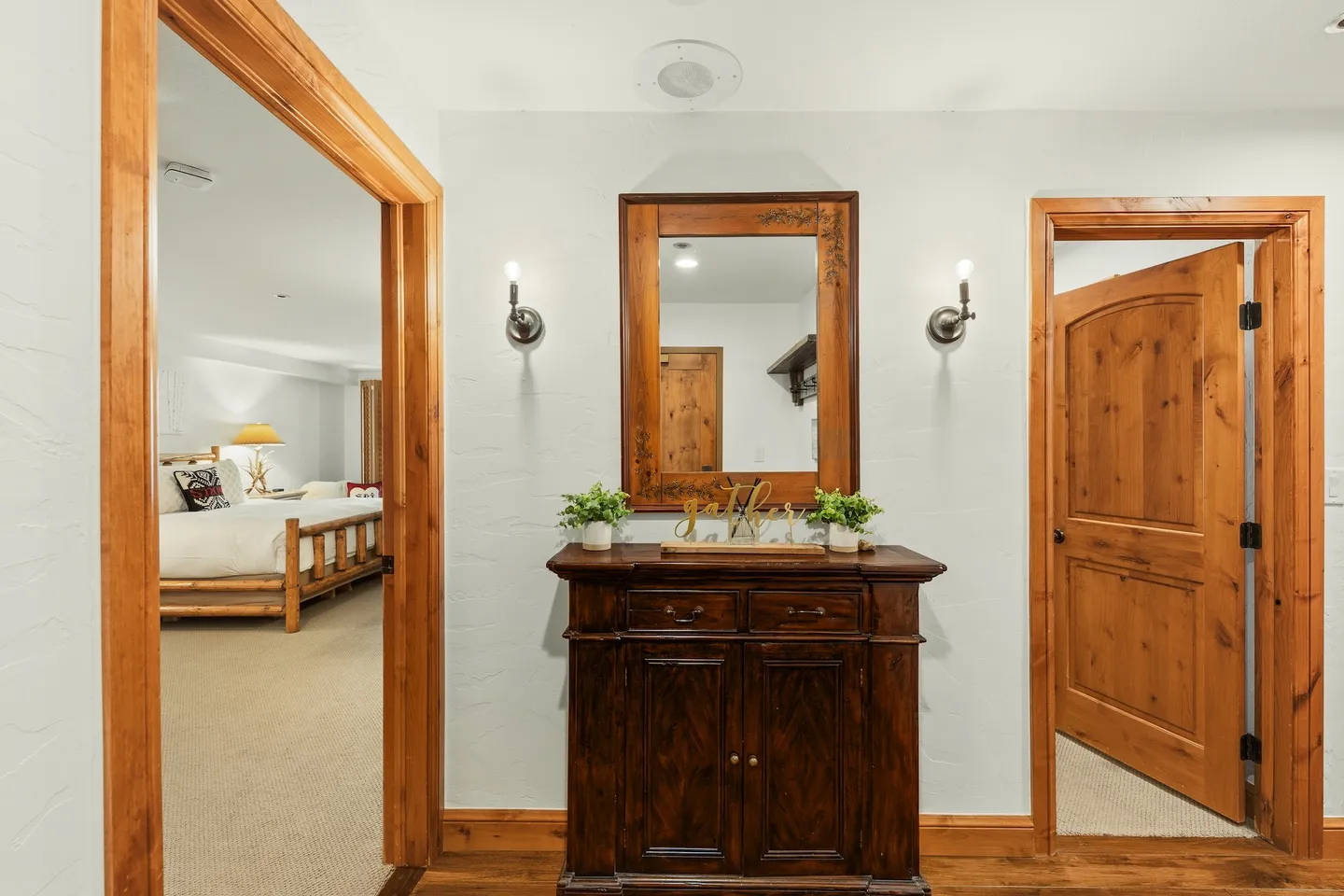 Hallway featuring a dark wood console cabinet beneath a framed mirror flanked by wall sconces, with doorways leading to an adjoining bedroom.