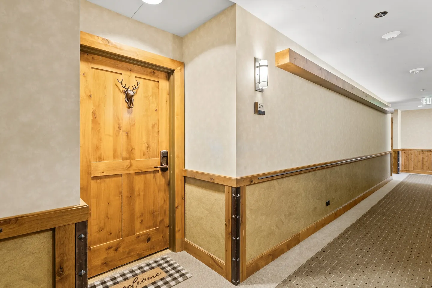 Carpeted hallway showing a wooden apartment entrance door with a deer head decoration and rustic wainscoting.