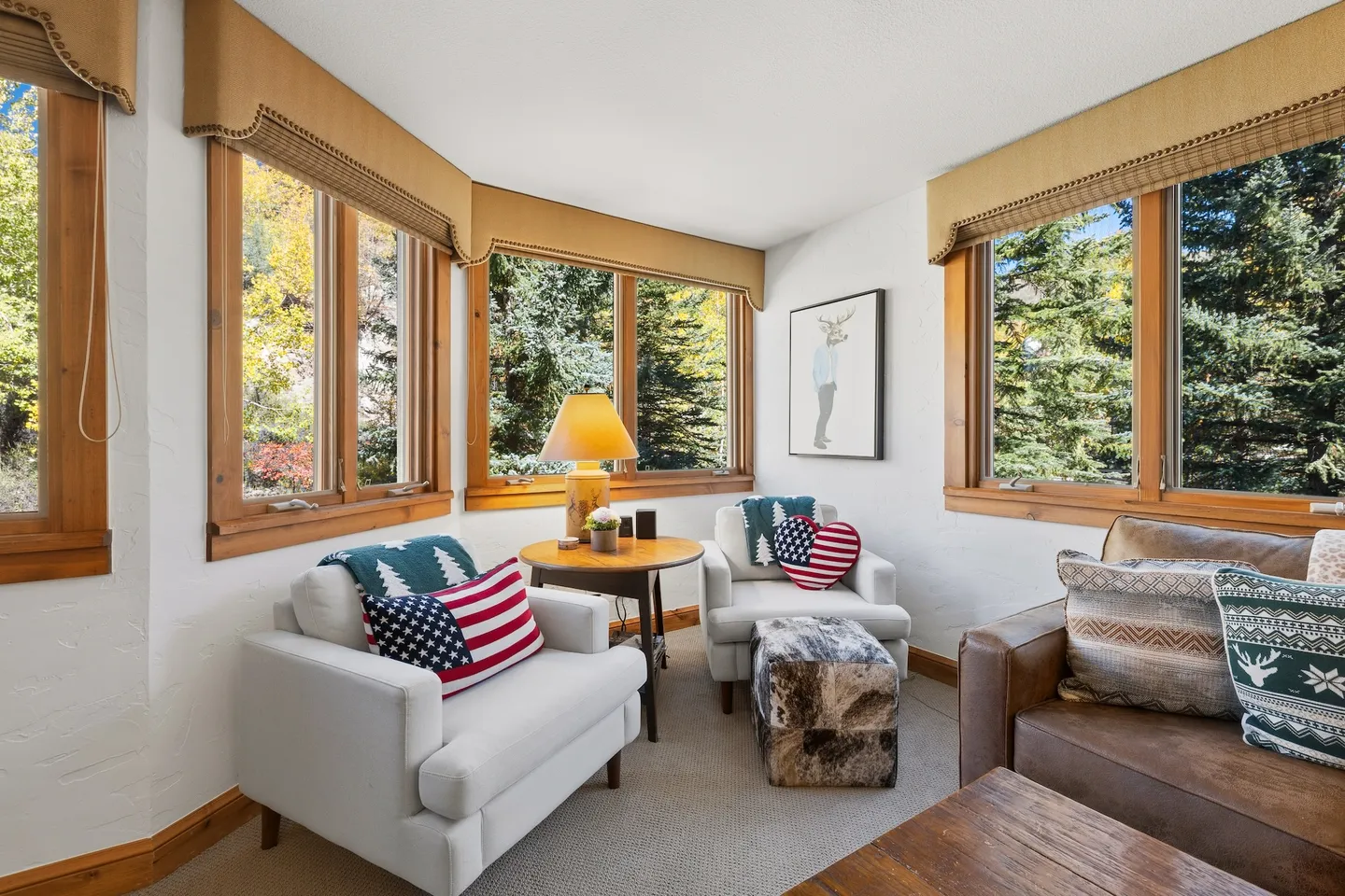 Sunlit sitting area with armchairs, an ottoman, a round end table with a lamp, and large windows looking out to pine trees.