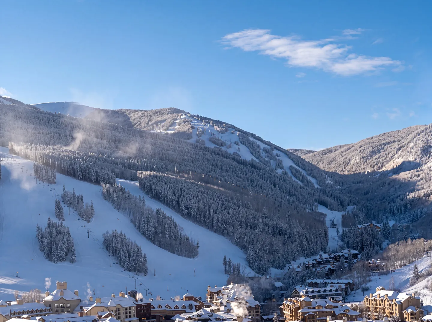 Elevated view of snow-covered ski slopes, pine forests, and village buildings under a clear blue sky.