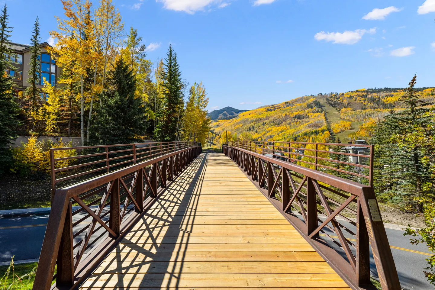 Wooden pedestrian bridge with metal railings leading toward mountain hills with autumn foliage under a blue sky.