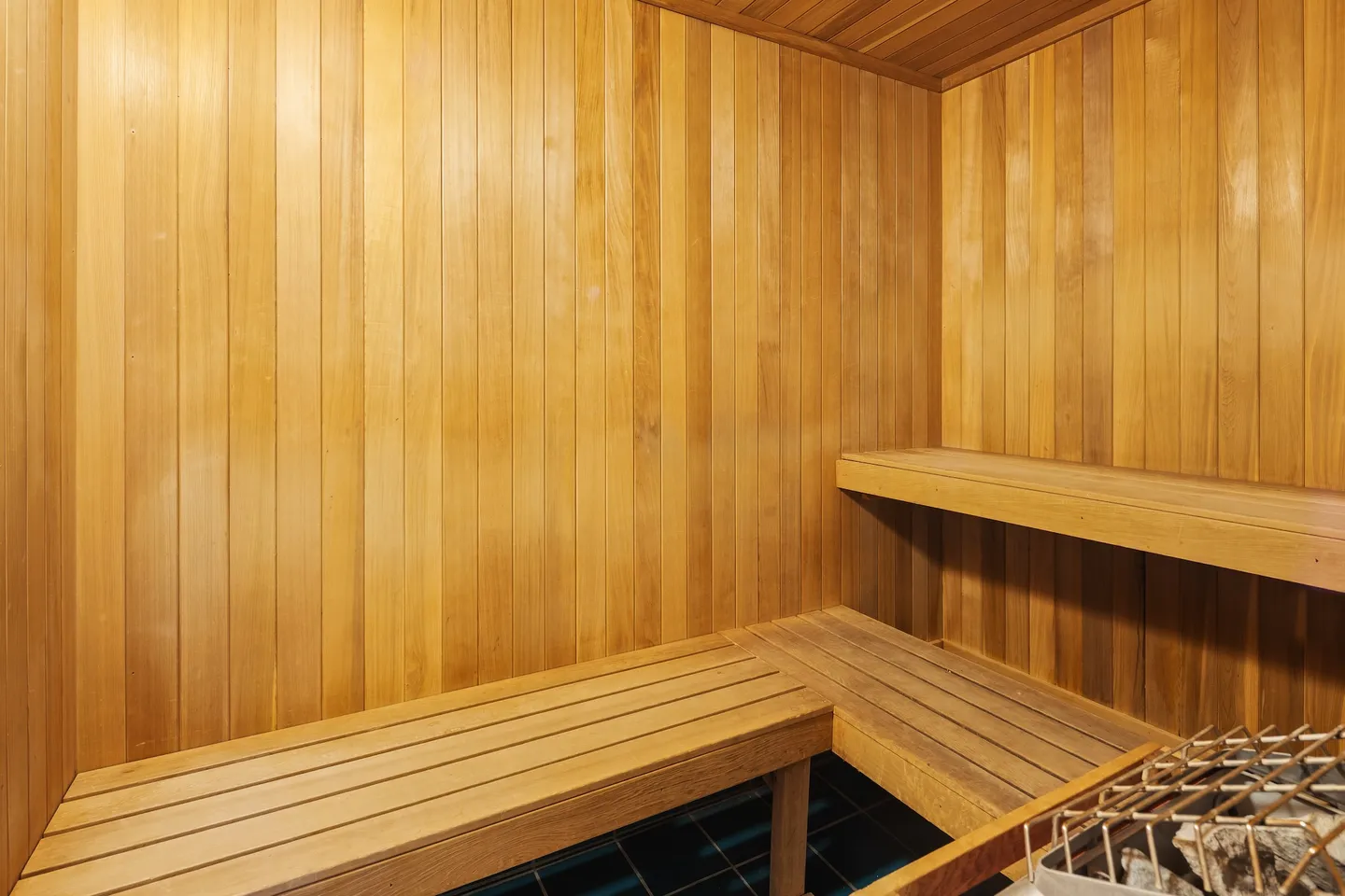 Indoor wood-paneled sauna featuring two-tiered slatted benches and a sauna heater filled with rocks in the foreground.