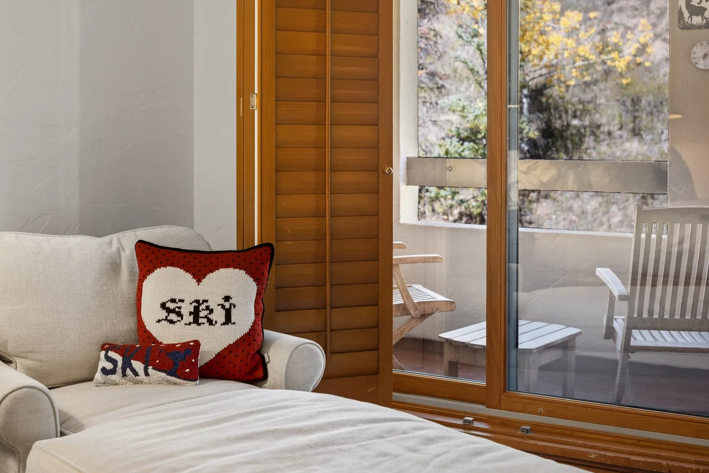 A chaise lounge with ski-themed throw pillows sits beside a sliding glass door leading to an outdoor balcony with seating.