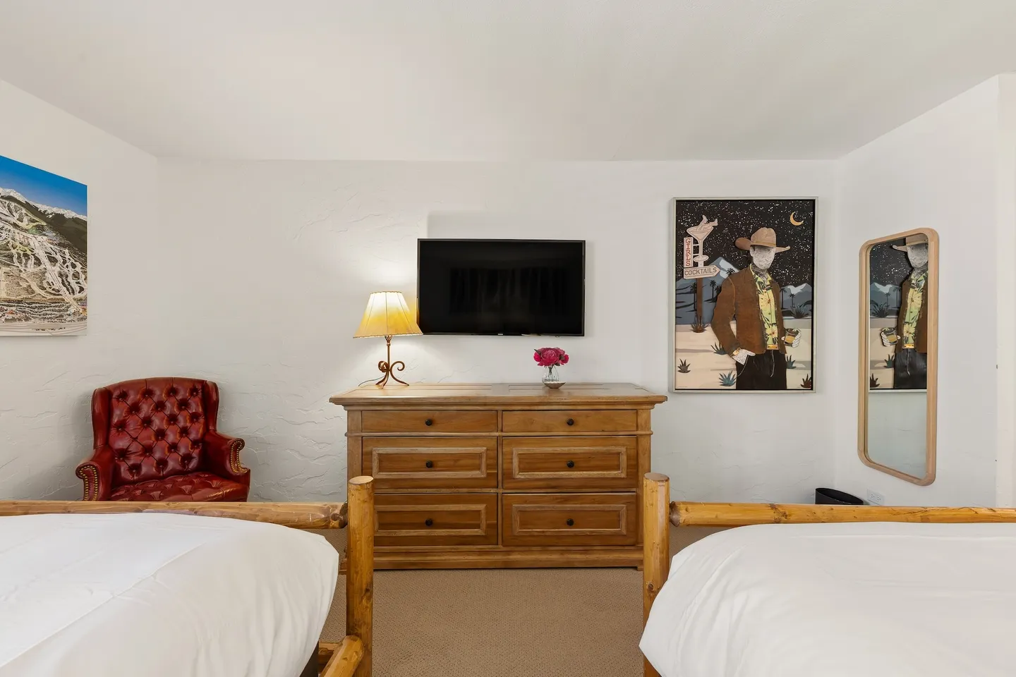 Bedroom featuring two beds in the foreground, a wooden dresser with a mounted TV, a red leather tufted armchair, and wall art.