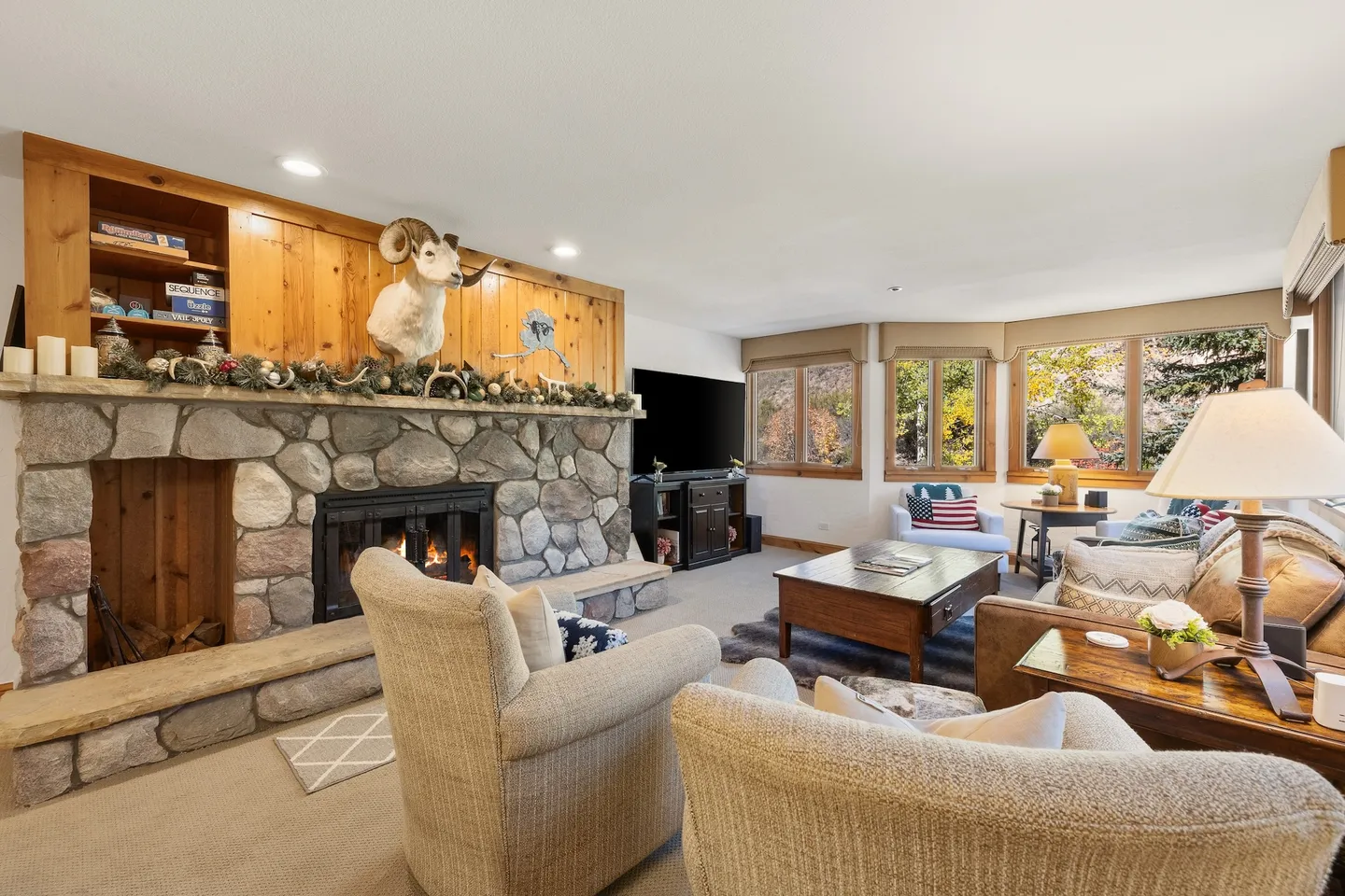 Living room with a large stone fireplace, mounted animal head, armchairs, sofa, and windows overlooking autumn foliage.