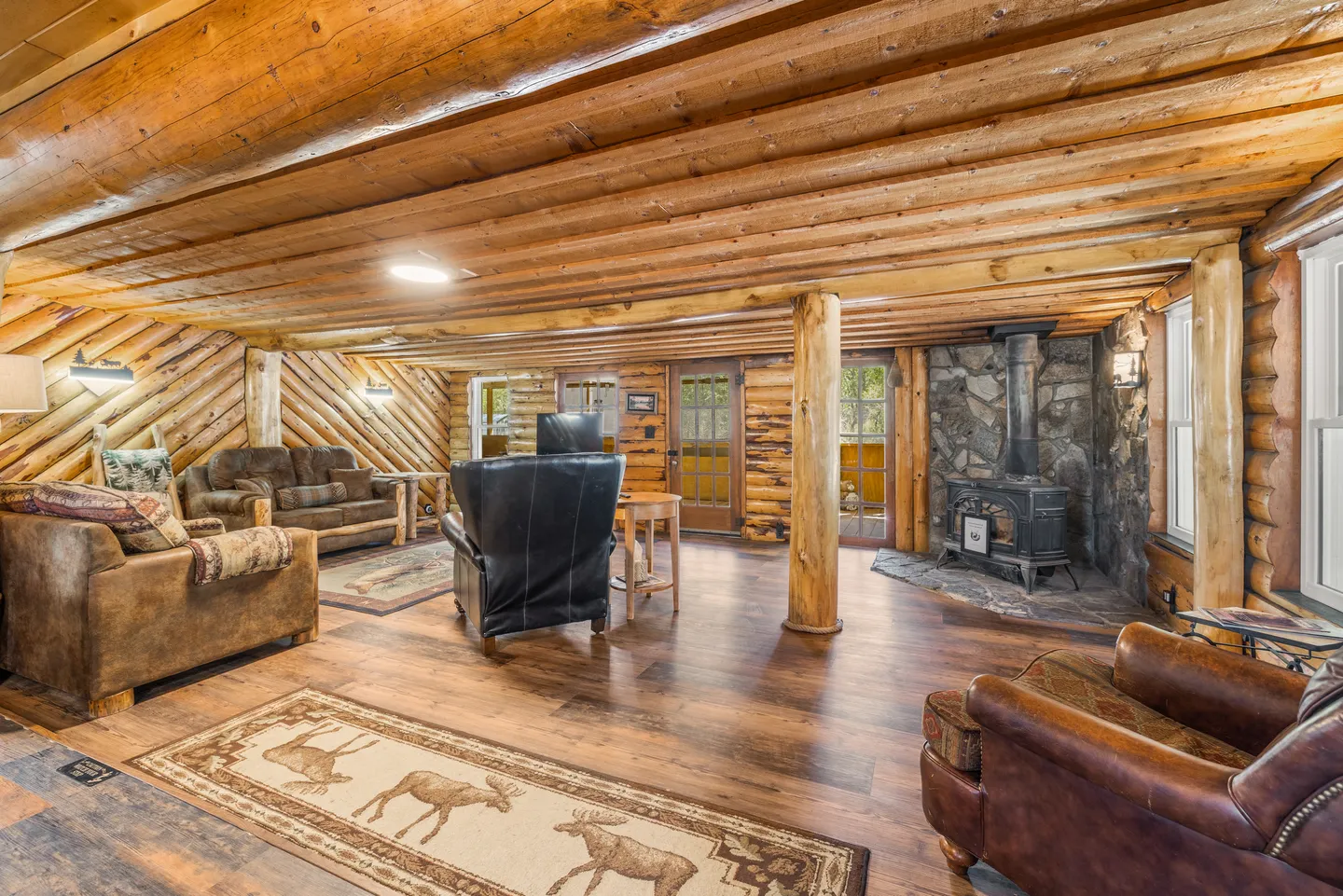 A rustic living room with log wood ceiling and walls, wood-look flooring, multiple armchairs, a couch, and a wood-burning stove on a stone hearth.