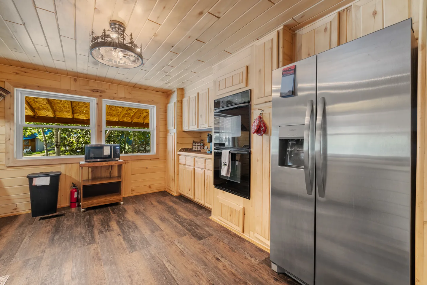 Kitchen with wood-paneled walls and ceiling, stainless steel refrigerator, built-in double oven, and two windows.