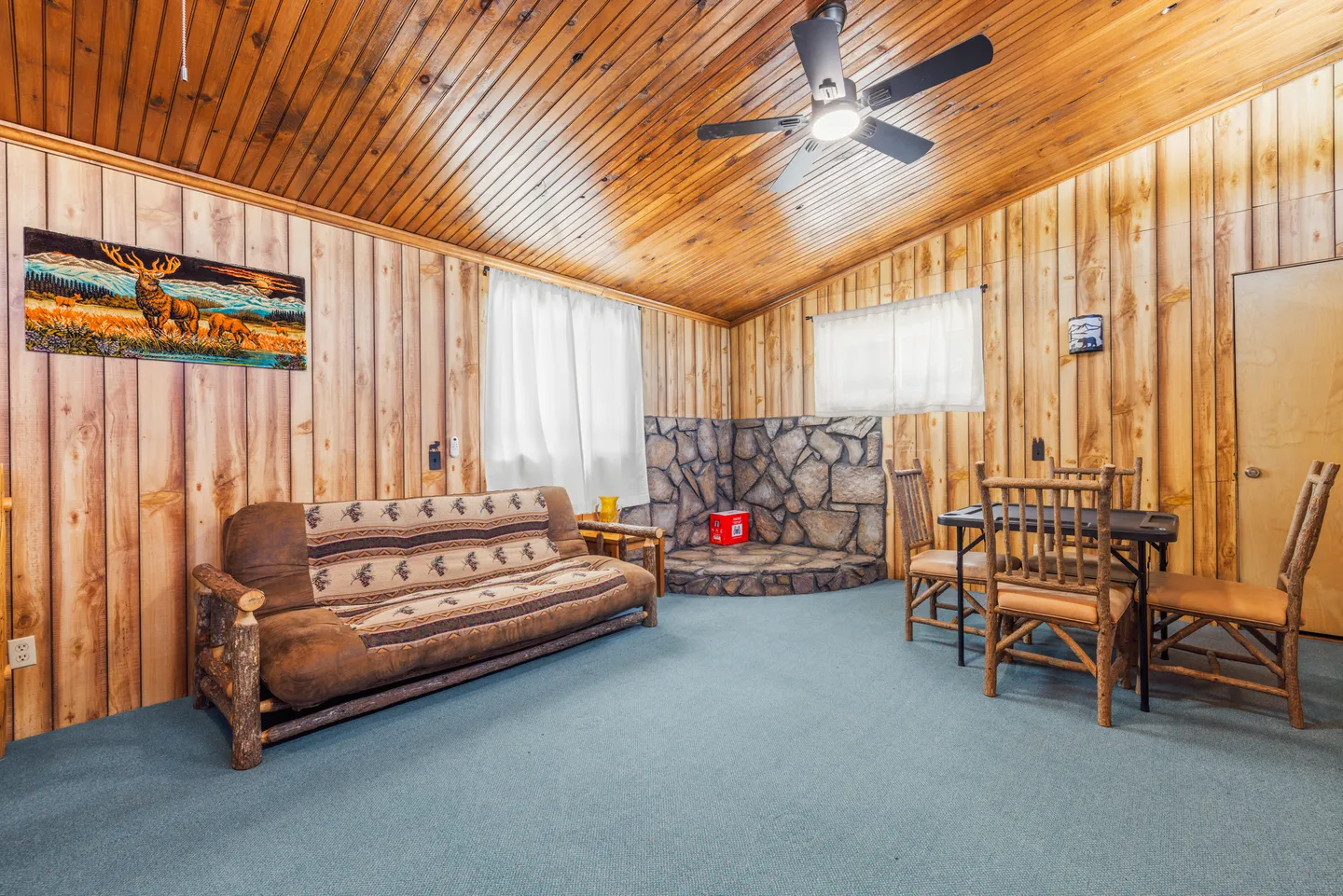 A living area with wood-paneled walls and ceiling, a futon sofa, a dining table with rustic chairs, and a stone hearth corner.