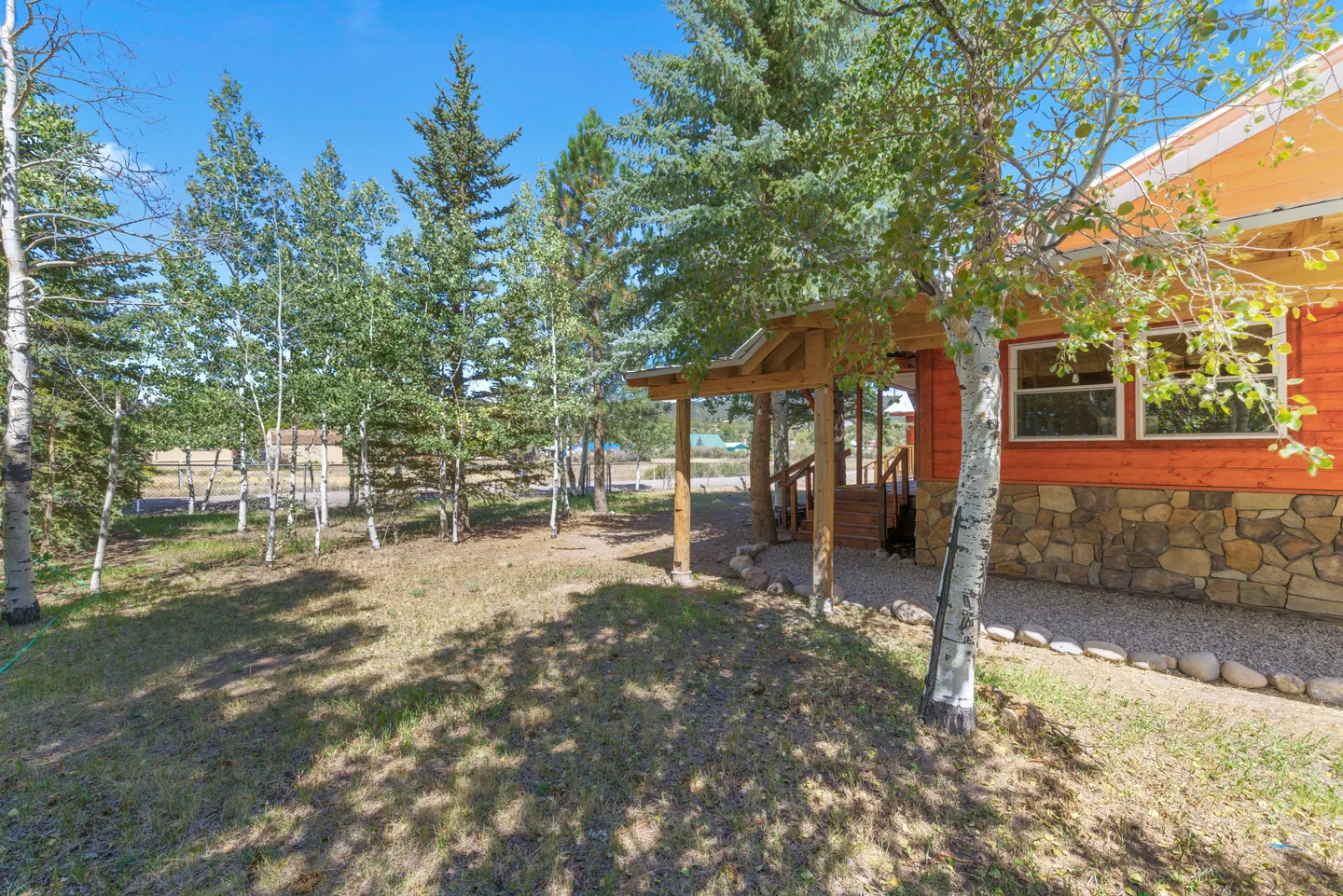 Exterior view of a home with orange wood siding, stone wainscoting, a covered porch, and a yard shaded by pine and birch trees under a clear blue sky.