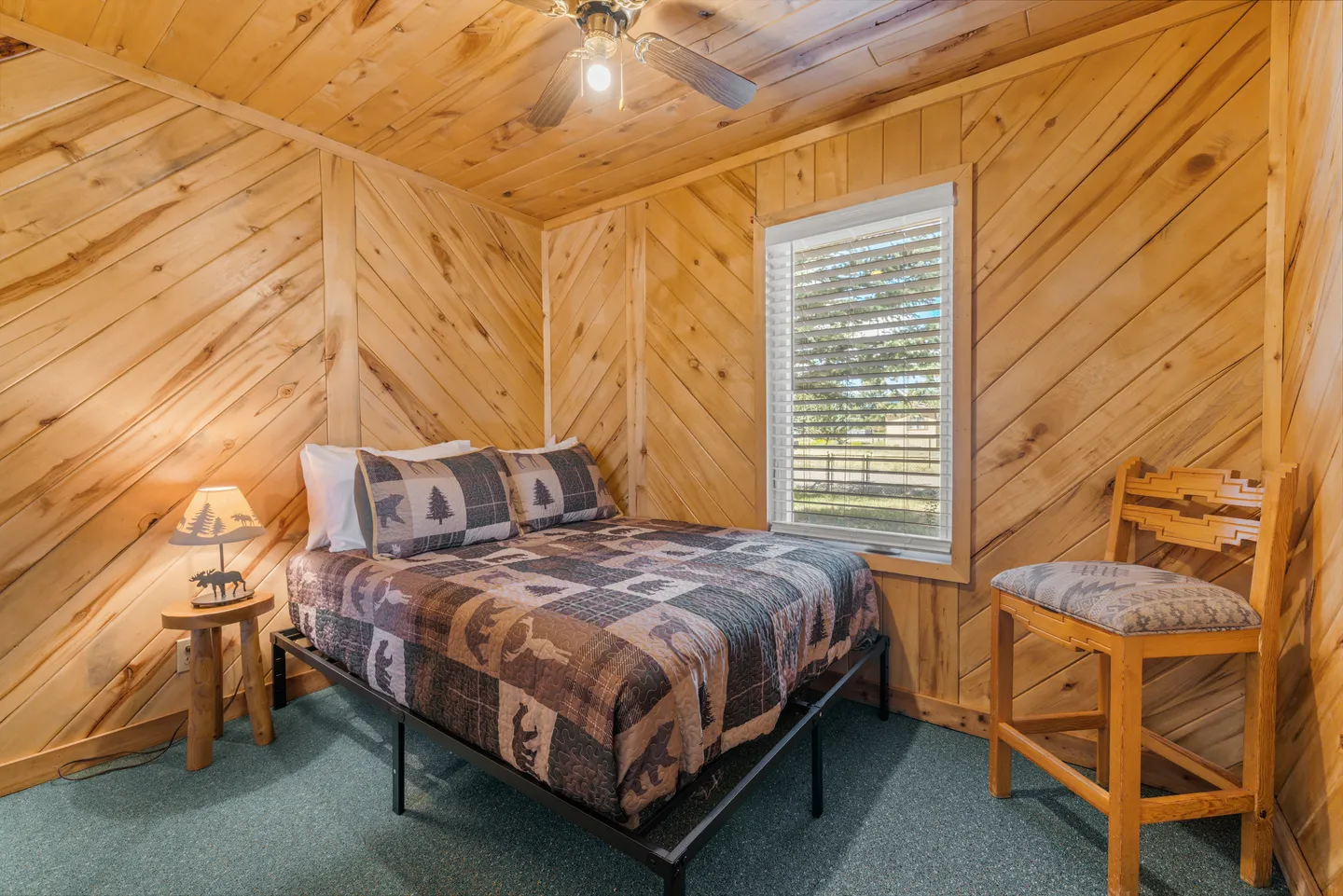Bedroom with diagonal wood-paneled walls and ceiling, green carpet, a bed with a rustic quilt, a wooden chair, and a window.