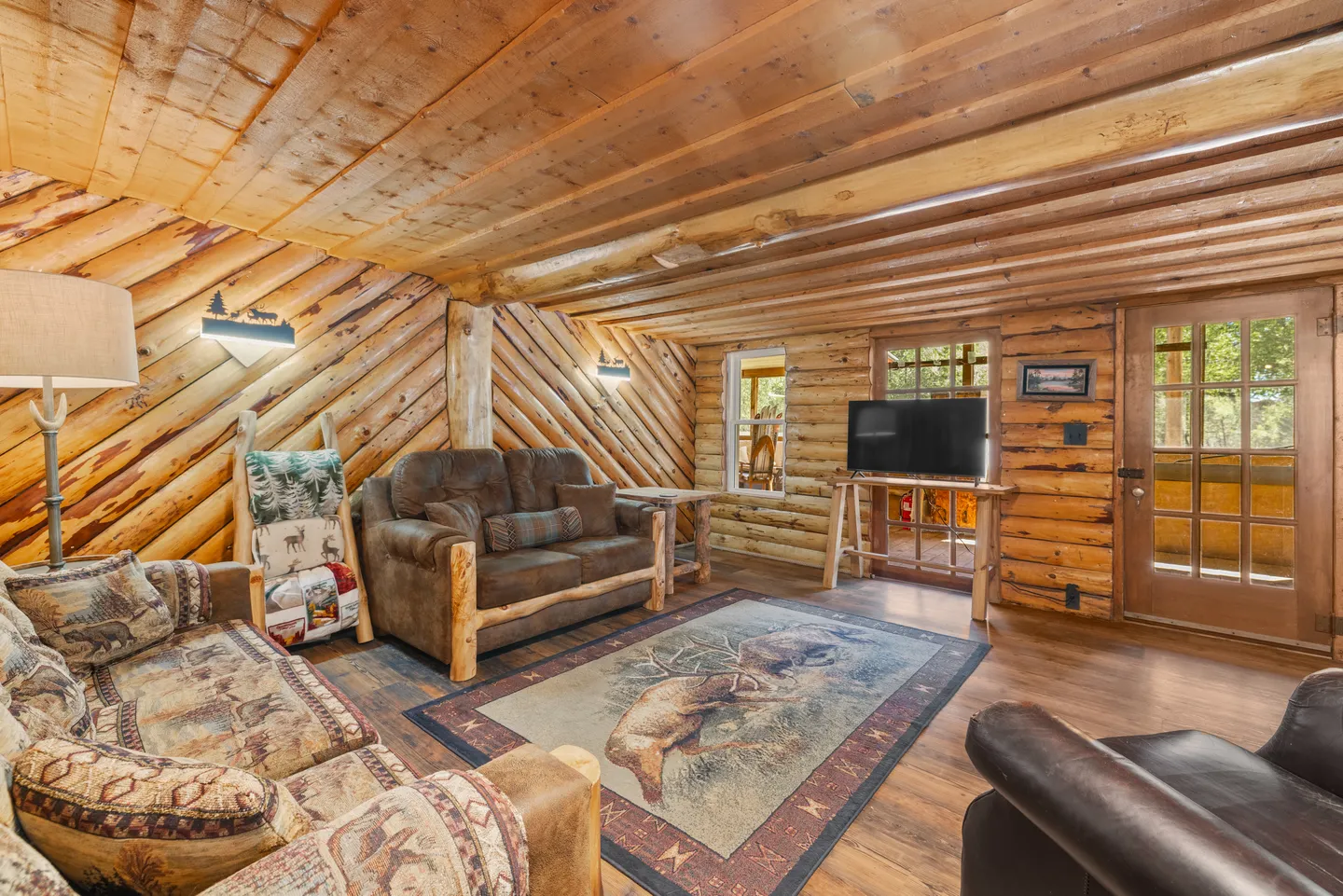 A rustic living room with log walls, wood-paneled ceiling, upholstered seating, and a television on a wood stand.