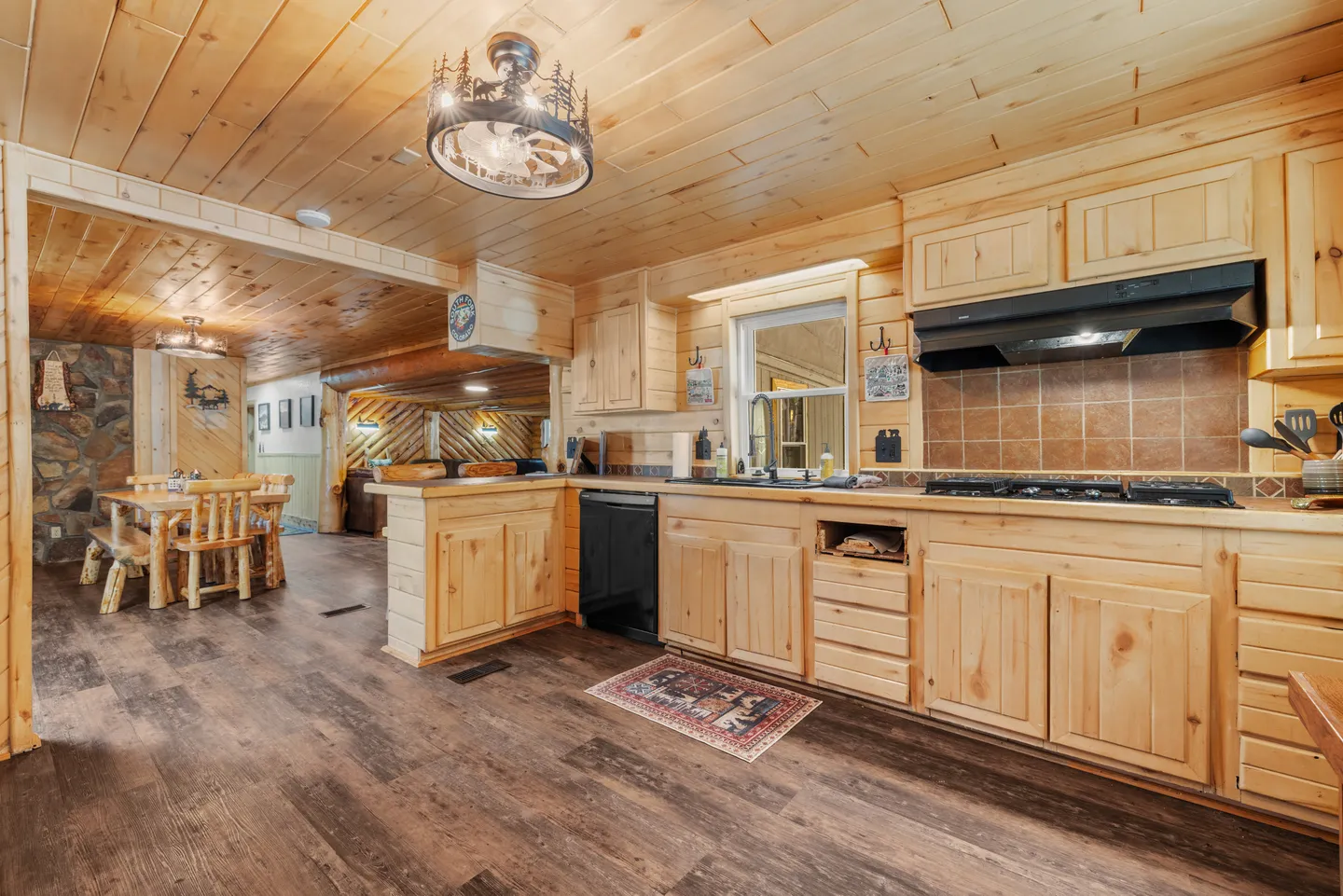 Open-concept cabin kitchen with pine wood cabinets, wood-paneled ceiling, dark flooring, and a view into the dining area.