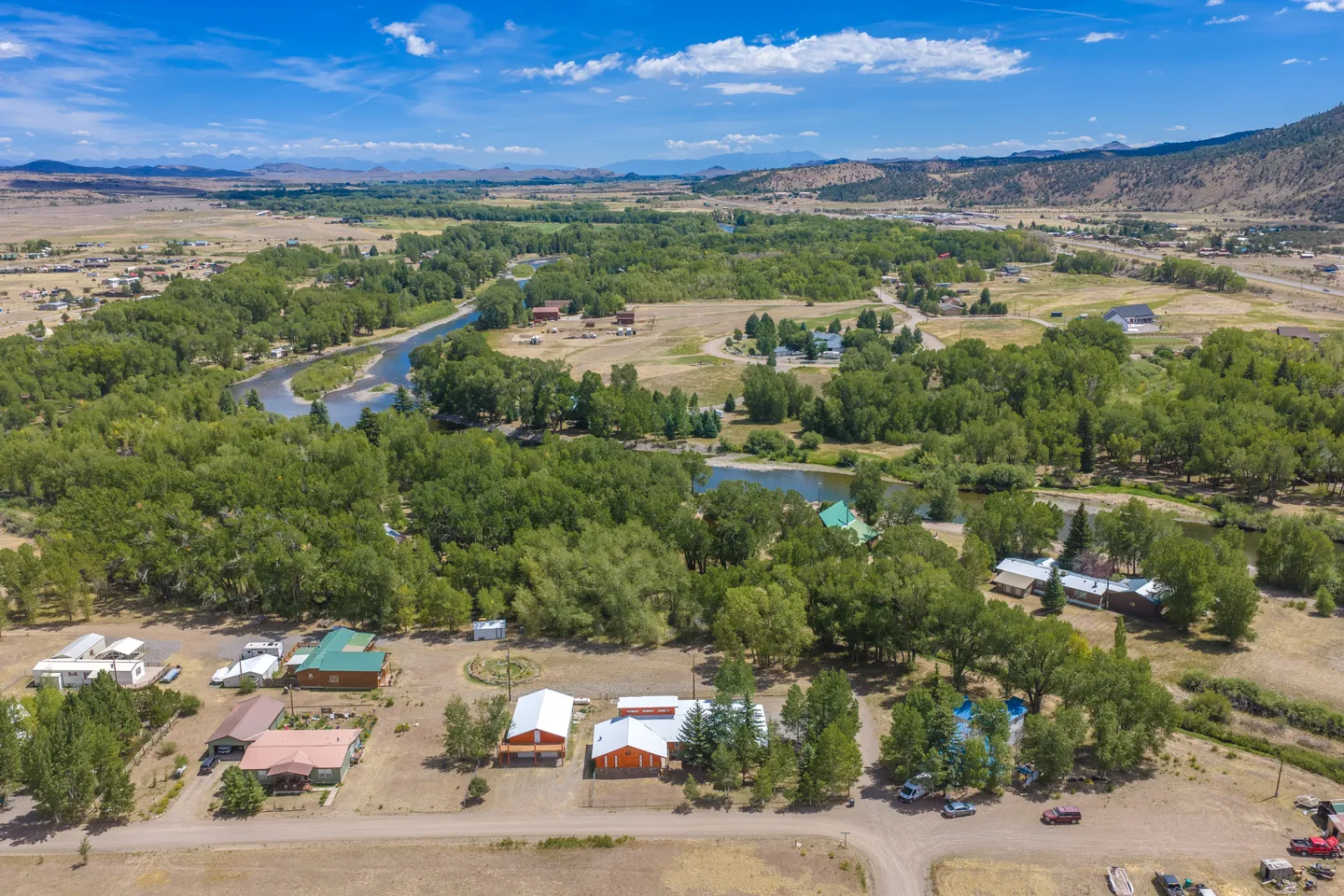 High aerial view of a rural valley with houses, a winding river, dense green trees, and distant mountains under a blue sky.