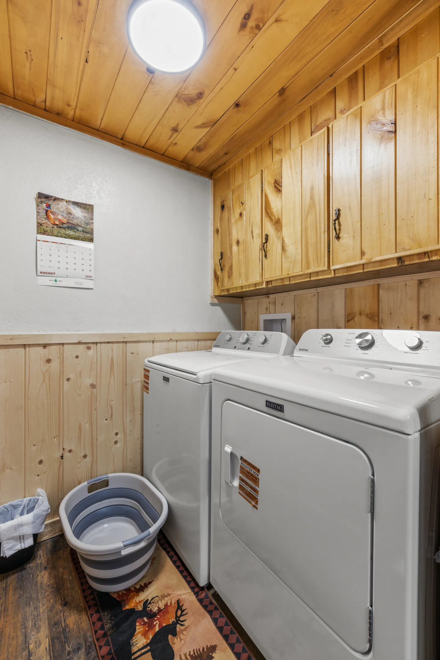 A laundry room with a white washing machine, dryer, wood cabinetry, and wood paneling on the walls and ceiling.