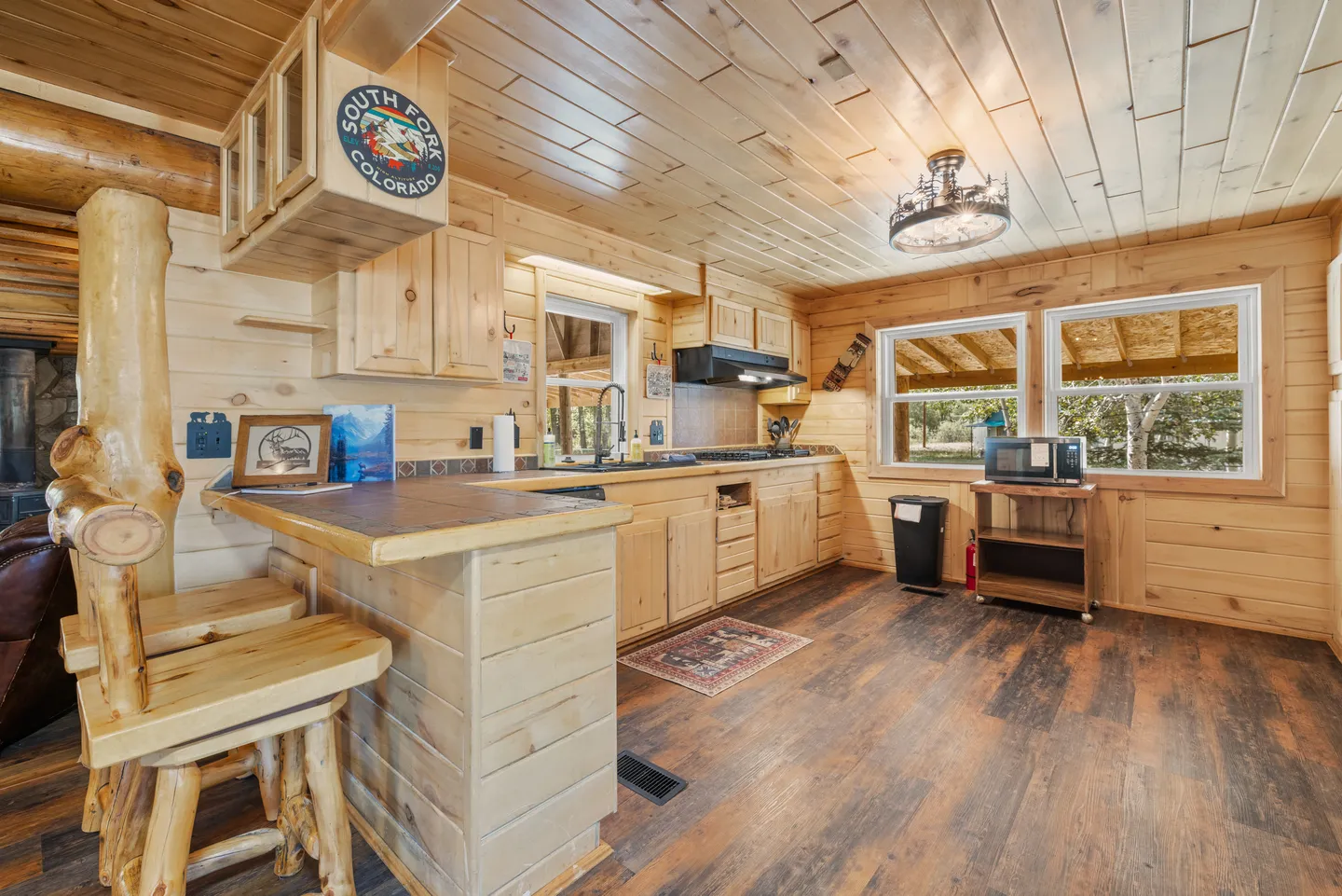 Cabin kitchen with knotty pine paneled walls and ceiling, wooden cabinets, a breakfast bar with log stools, and windows looking outside.