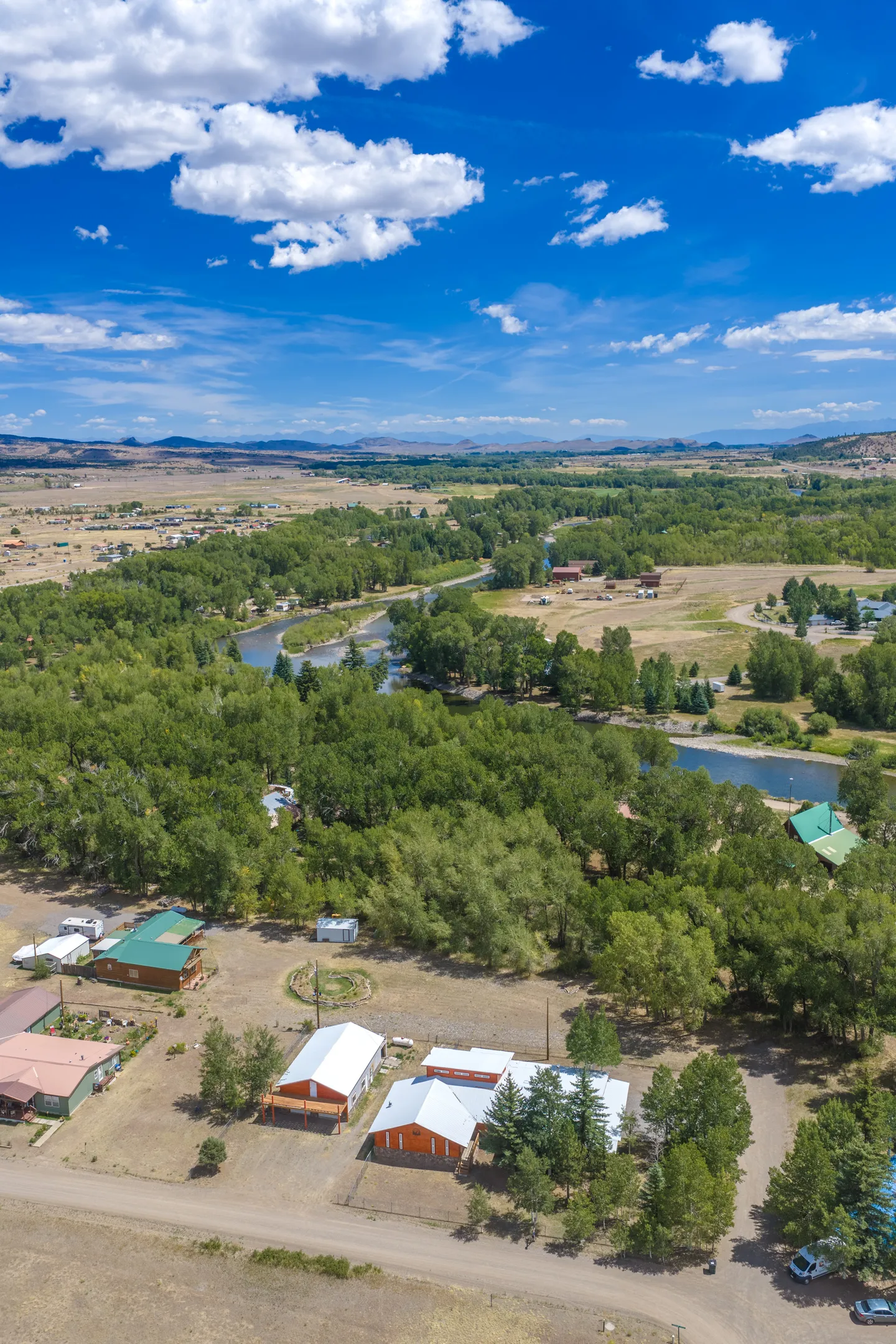 High aerial view of a river valley with lush green trees, scattered houses, and distant mountains beneath a bright blue sky.