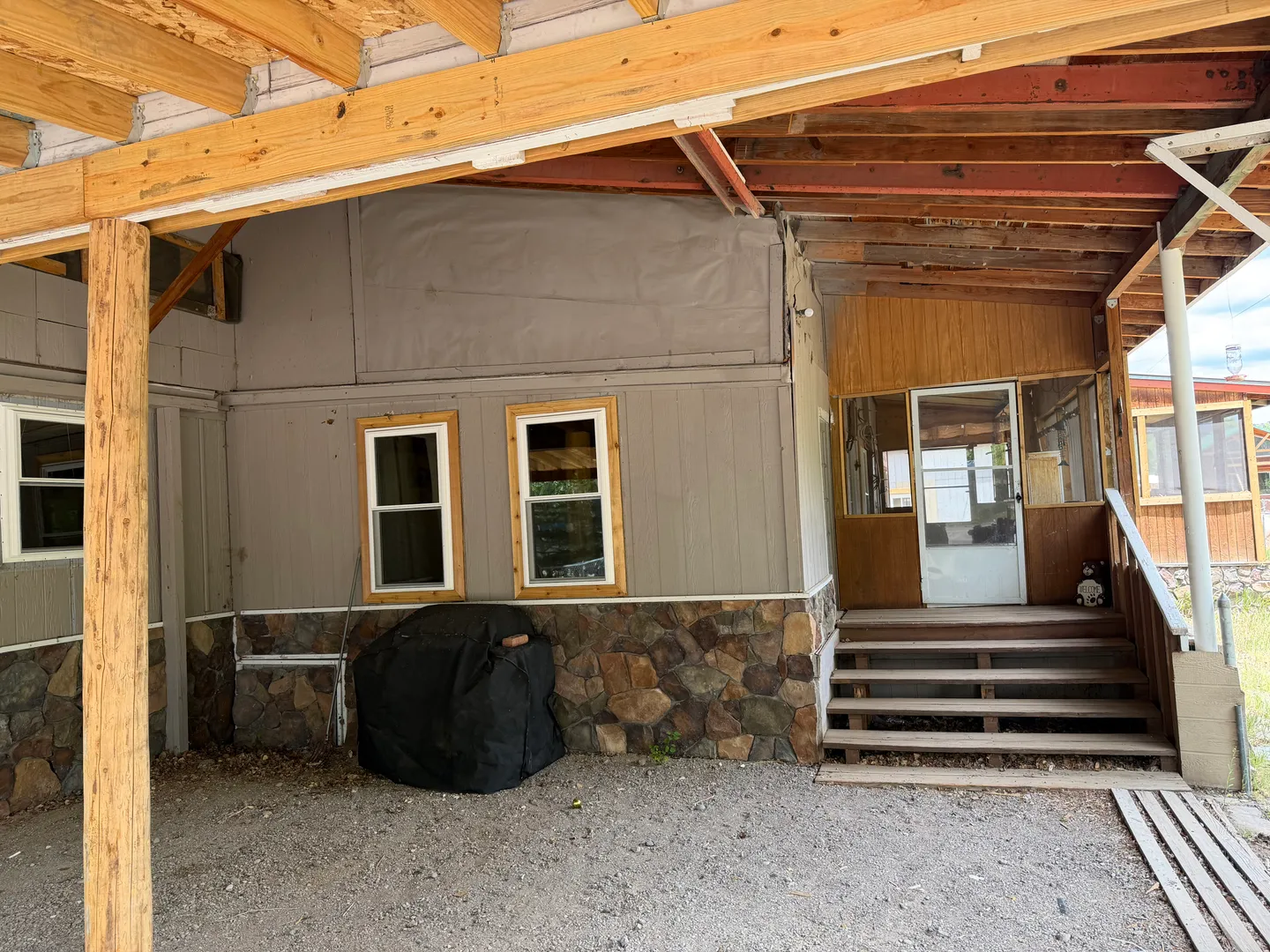 Exterior view of a home entrance underneath a covered wooden carport with stone wainscoting and wooden stairs.
