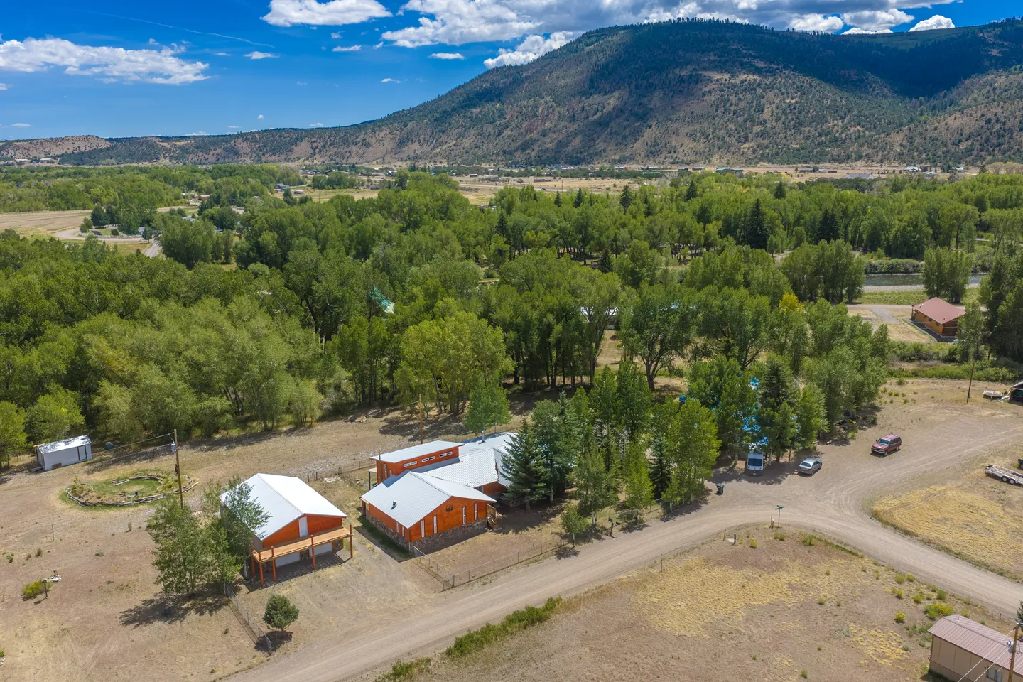 Aerial view of a rural property featuring orange buildings with metal roofs, surrounded by dense green trees and a mountain in the background.