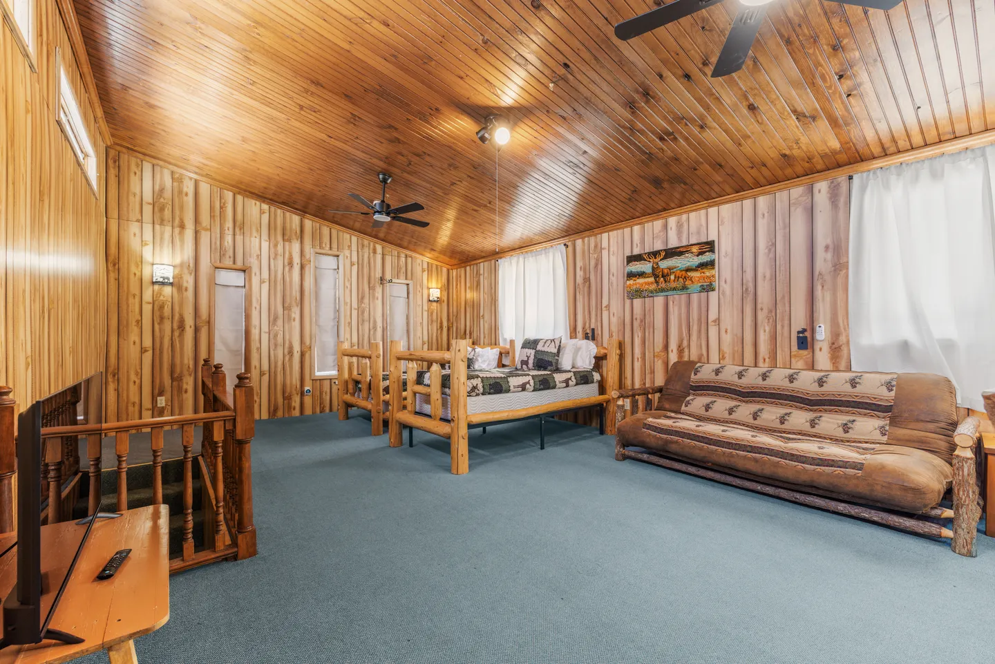 A rustic wood-paneled upper-level room with blue carpeting, log-frame daybeds, a log-frame futon, and a vaulted tongue-and-groove ceiling.