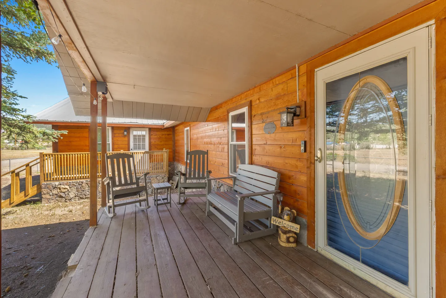 Front porch of a log-style cabin with wooden plank flooring, outdoor rocking chairs, a bench, and a glass-paneled front door.