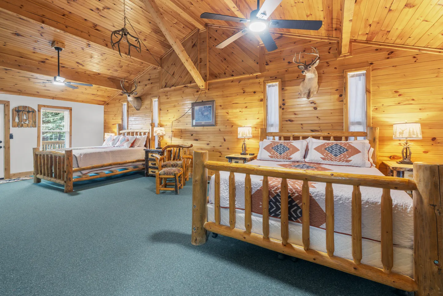 Spacious bedroom with wood-paneled walls and ceiling, two log frame beds, teal carpeting, and taxidermy mounts.