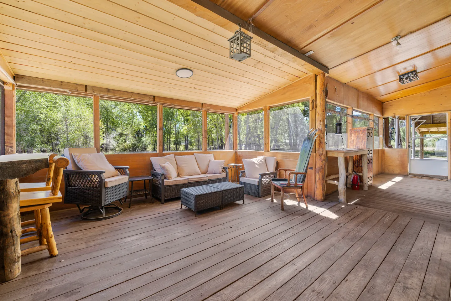 Screened porch with wood plank flooring, wood-paneled ceiling, and cushioned wicker patio furniture looking out at trees.