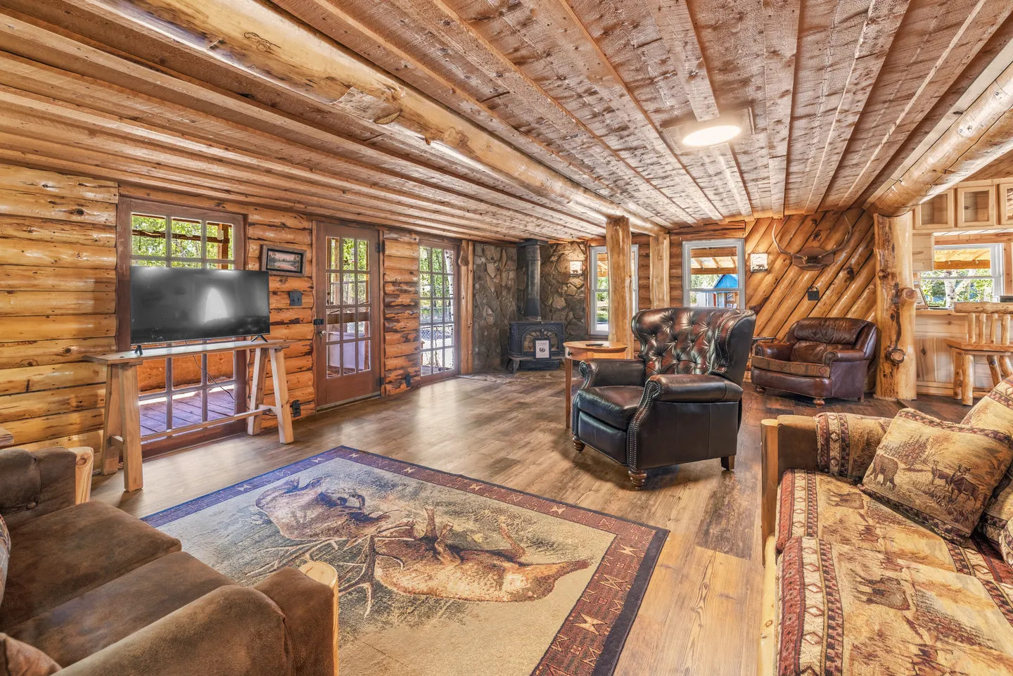 Rustic living room with log walls, wood-plank ceiling, leather armchairs, patterned couch, and a wood stove in the corner.