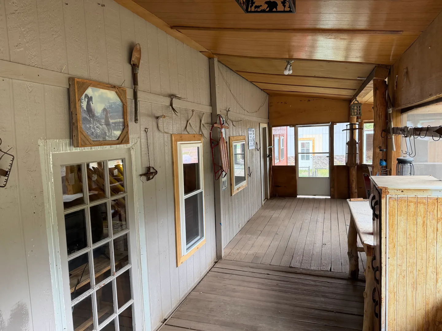 A long covered porch with wooden plank flooring, wood-paneled ceiling, and rustic decorative items hung on the siding.