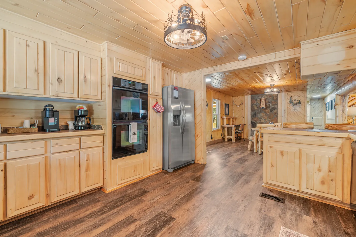 Kitchen with light wood cabinetry, double wall oven, stainless steel refrigerator, and wood-paneled walls and ceiling opening to a dining area.