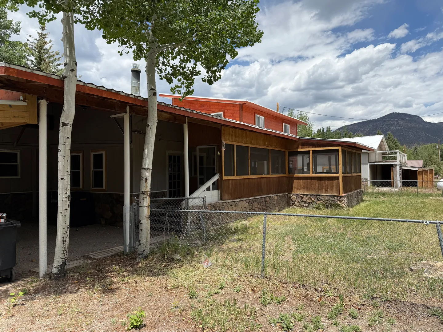 Exterior of a two-story home featuring a covered carport, screened porch, chain-link fence, and yard against a mountainous backdrop.