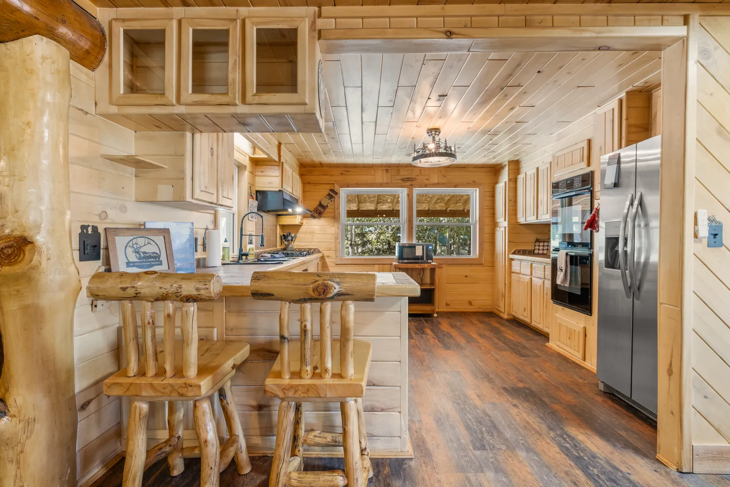 Rustic cabin kitchen with light pine paneling, log barstools, stainless steel appliances, and wood-look flooring.