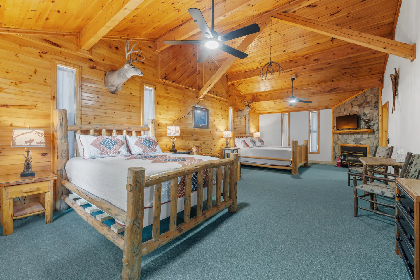 Spacious log cabin-style bedroom featuring wood-paneled walls and ceilings, two log-frame beds, and a stone fireplace.