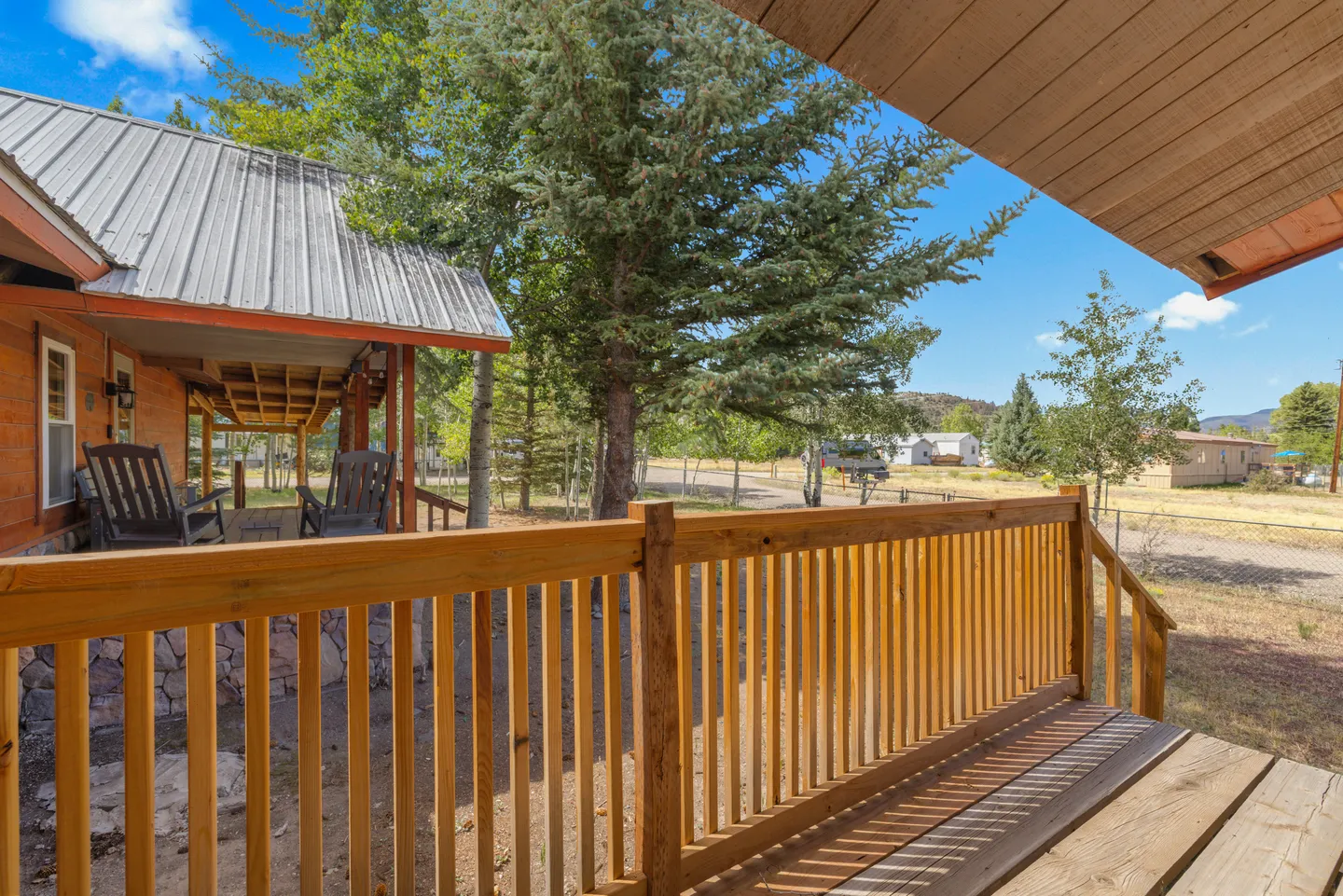 View from a wooden porch looking out over a front yard with trees toward a neighboring cabin and street.