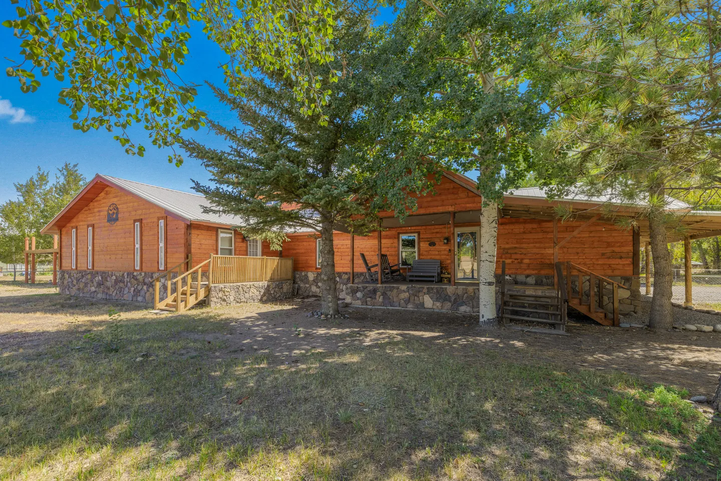 Exterior view of a log-style cabin with a stone foundation, covered front porch, and surrounding shade trees under a clear blue sky.