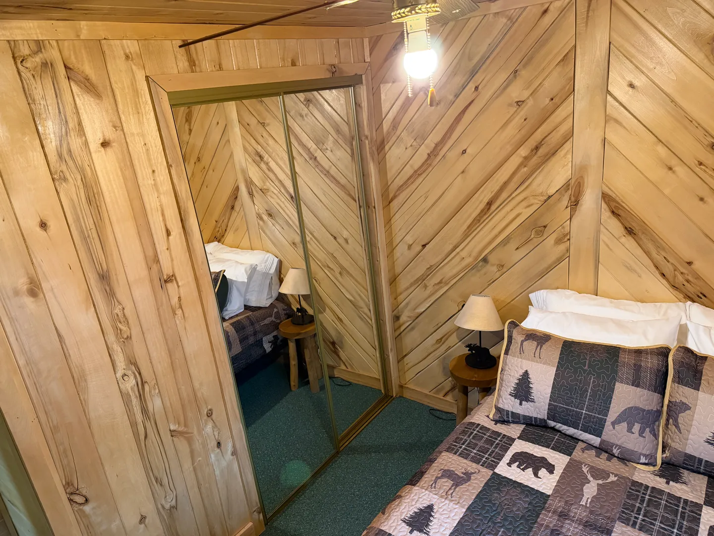 Bedroom with diagonal wood-paneled walls, carpeted flooring, a bed with a rustic quilt, and mirrored sliding closet doors.