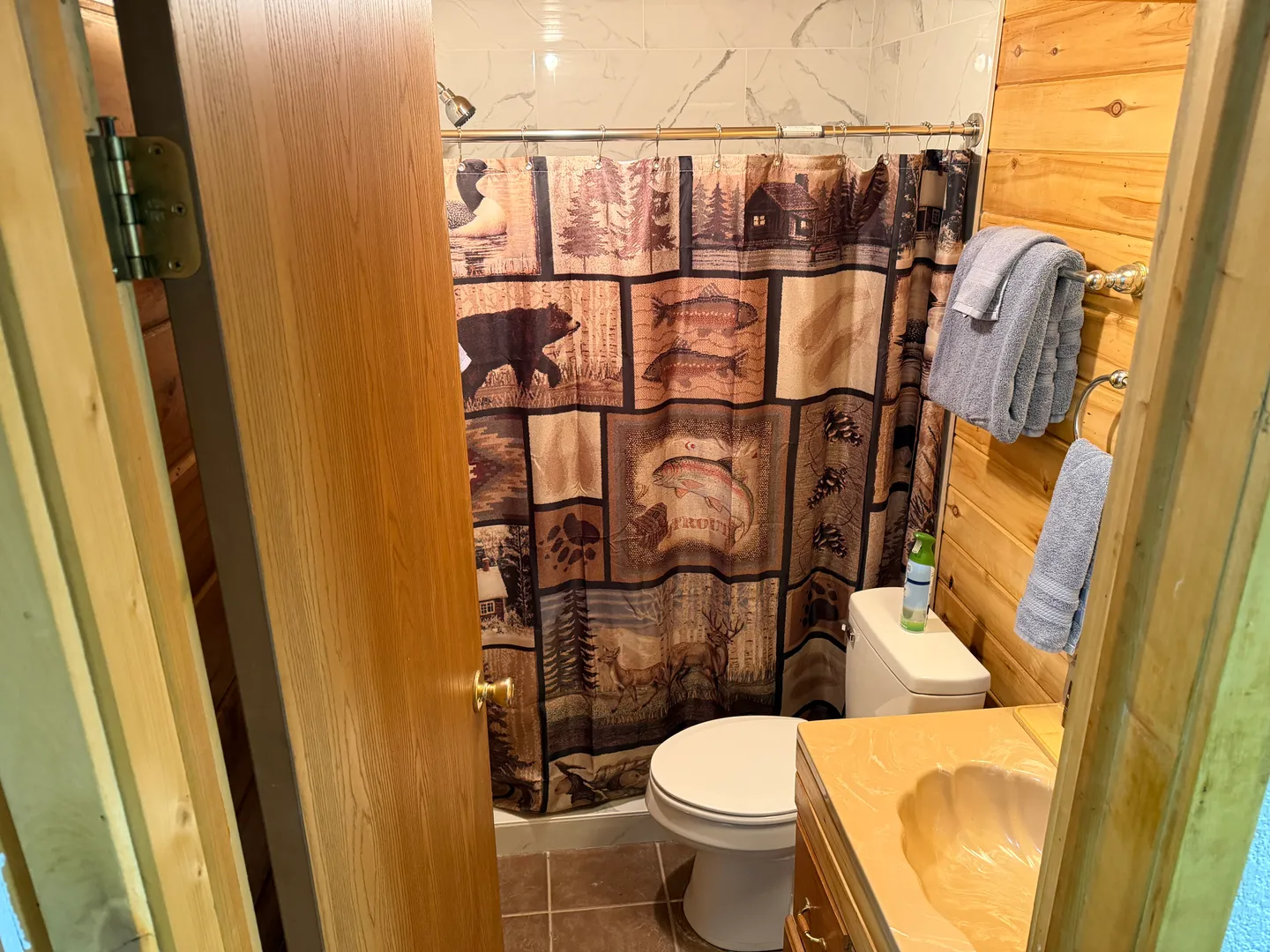 Bathroom with wood-paneled walls, a rustic wildlife shower curtain, toilet, and vanity with a shell-shaped sink.