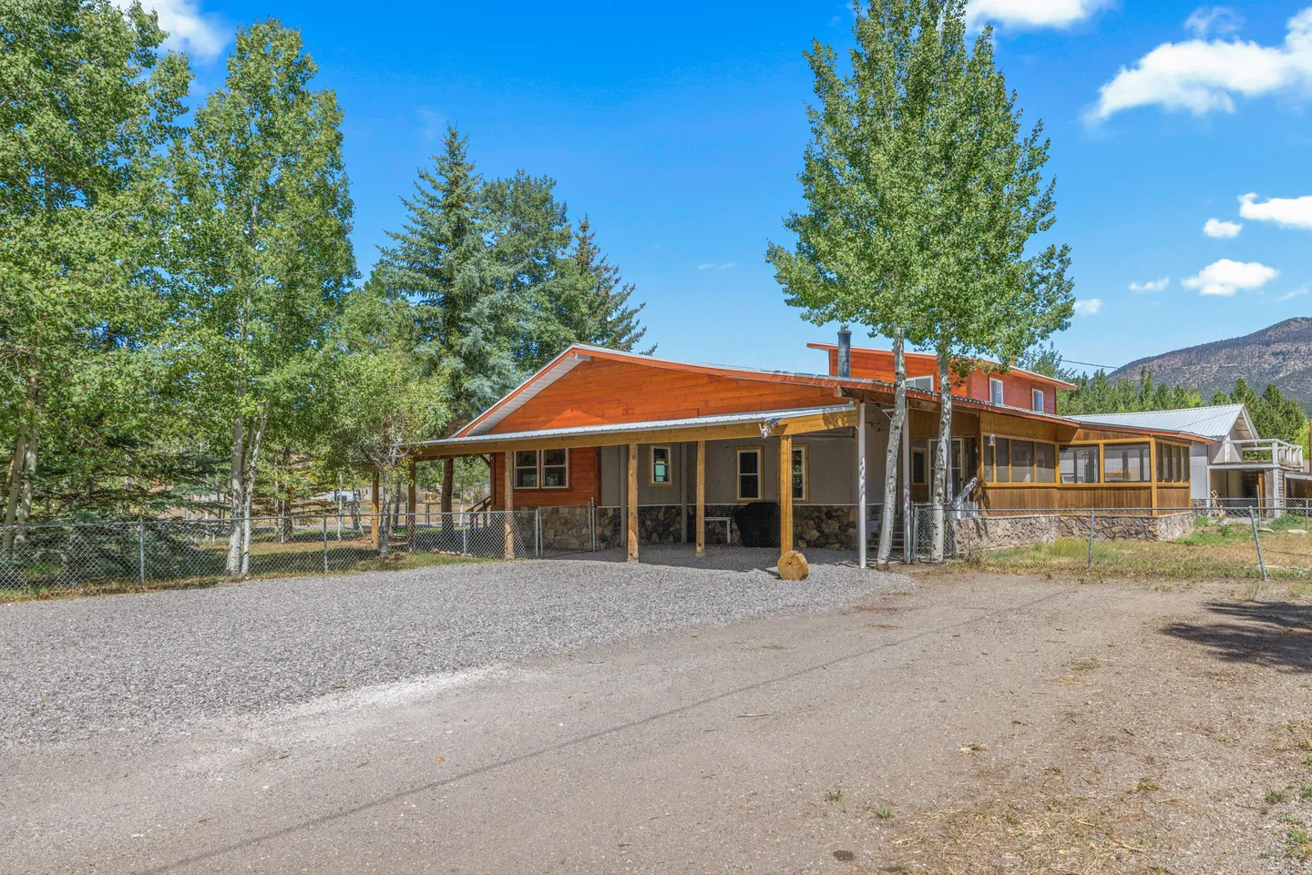 Exterior of a wood and stone house with a carport, screened porch, gravel driveway, and tall trees under a blue sky.